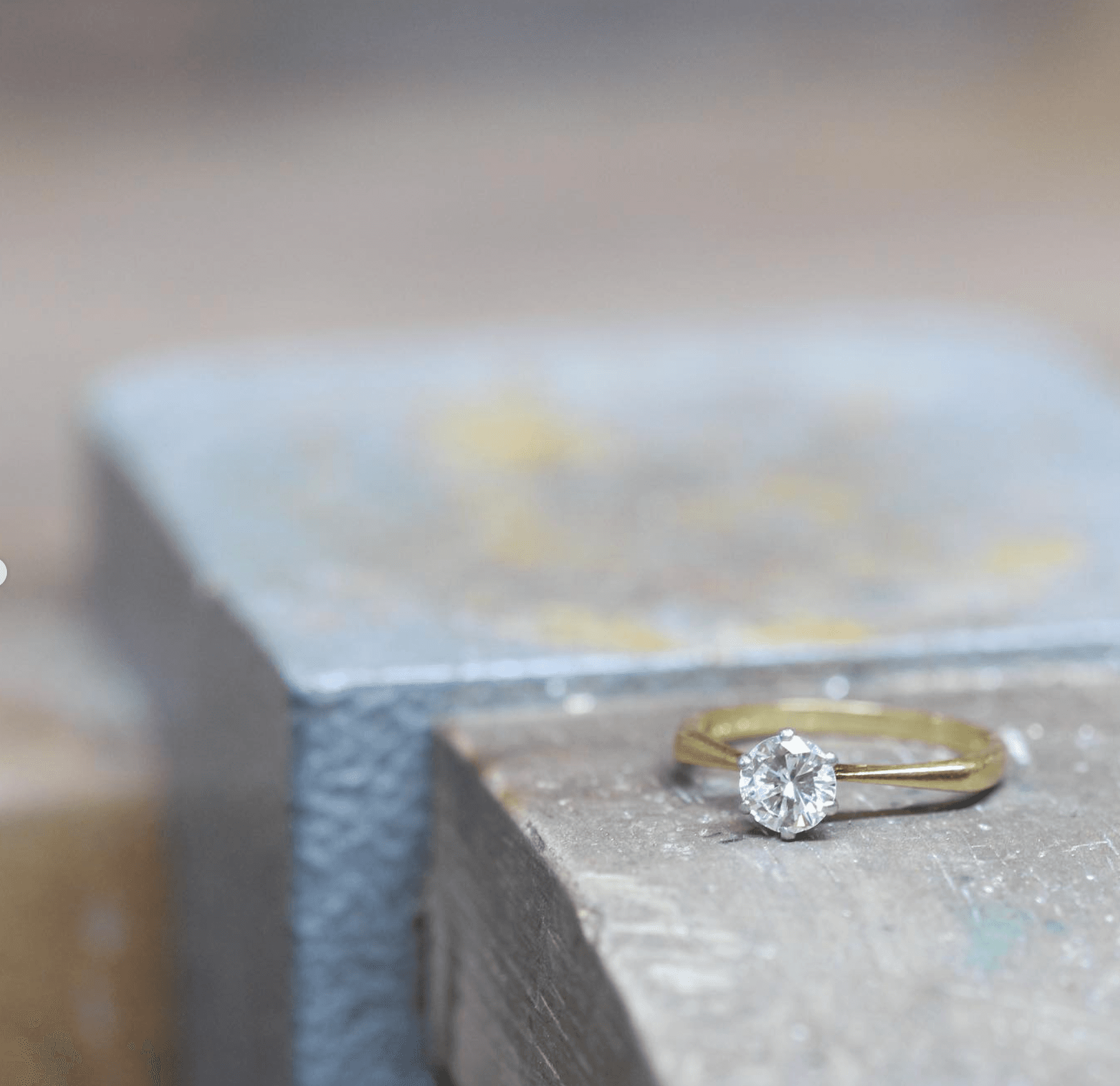 Freshly finished gold solitaire diamond engagement ring resting on the corner of a worn steel workshop anvil, with the London goldsmithing studio visible and soft bokeh behind