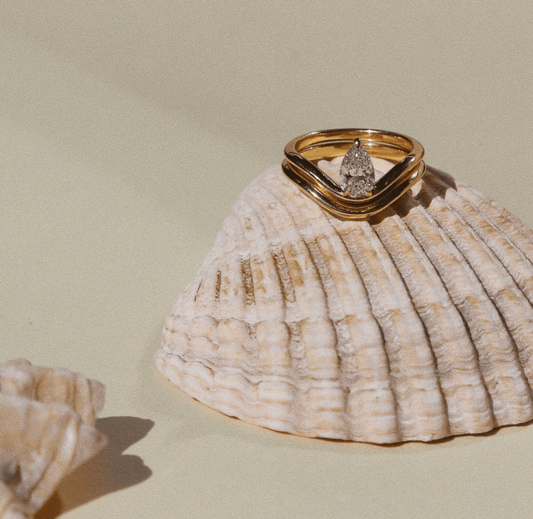 Pear-cut diamond chevron engagement ring stacked with matching curved wedding band, resting on a ridged sea shell against a warm sand background