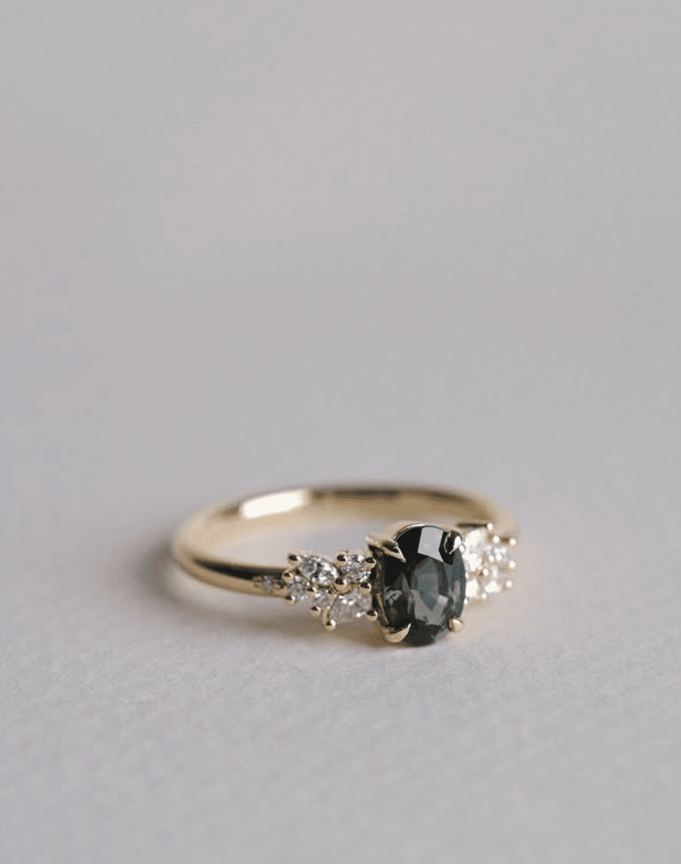 Teal oval sapphire engagement ring with diamond clusters shoulder-set either side, in 18ct yellow gold, against a warm grey studio background
