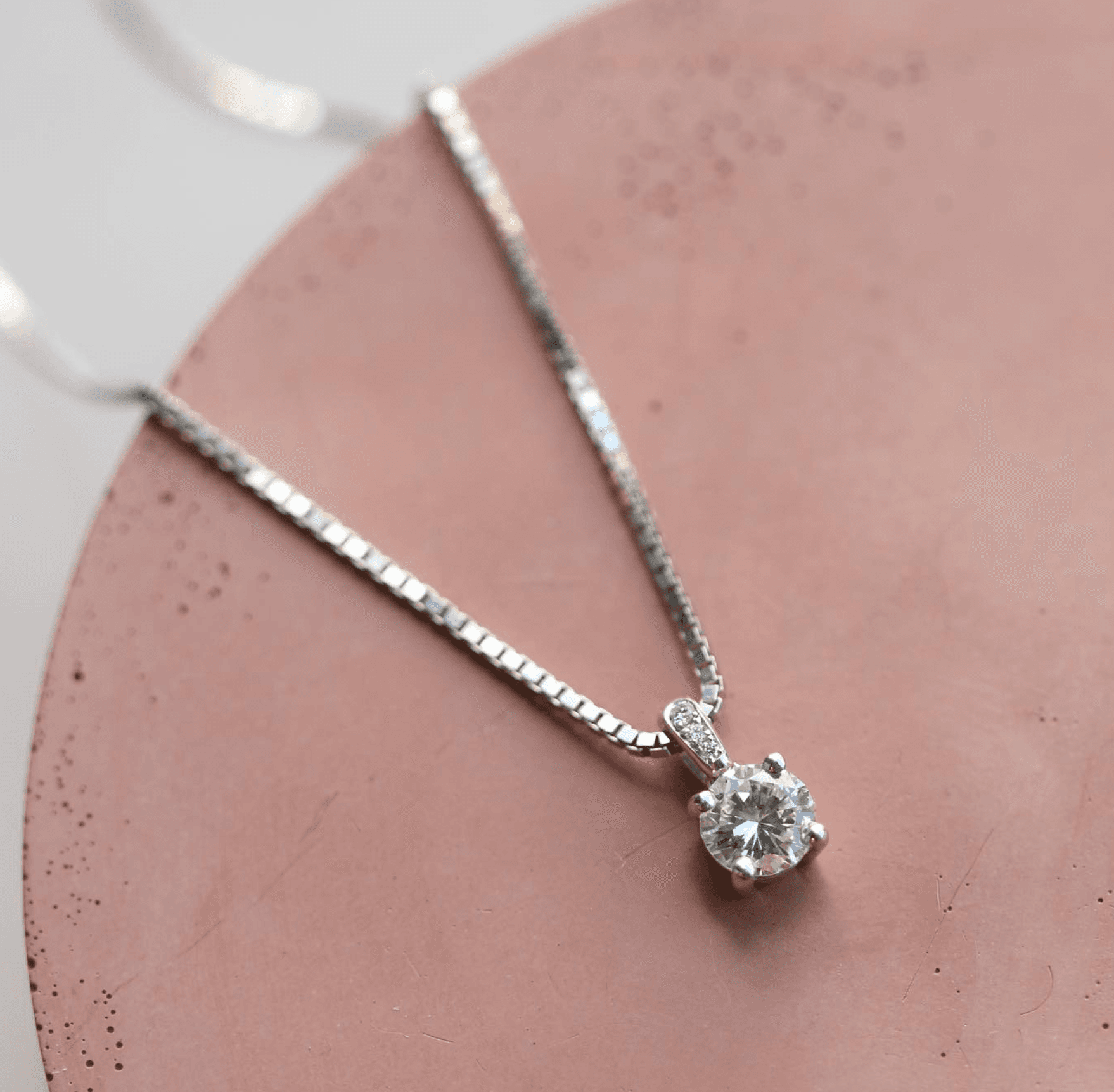 Round brilliant diamond solitaire pendant on a fine silver box chain, photographed on a blush pink surface